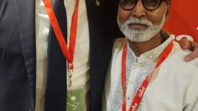 Two men posing for a photo at a conference: one in a dark suit with a red lanyard, the other in a white traditional outfit with glasses, both smiling in front of a red backdrop and wearing event badges.