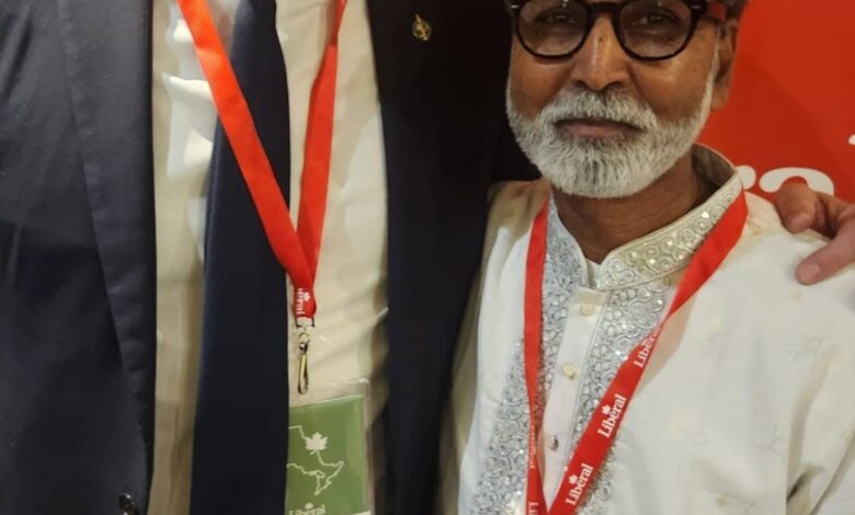 Two men posing for a photo at a conference: one in a dark suit with a red lanyard, the other in a white traditional outfit with glasses, both smiling in front of a red backdrop and wearing event badges.