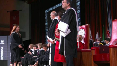 Graduate in a cap and gown walks across the stage to receive a diploma as faculty in robes stand and sit nearby on the platform.