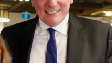 Smiling man in a dark suit and blue tie at an indoor event with warm lighting in the background.