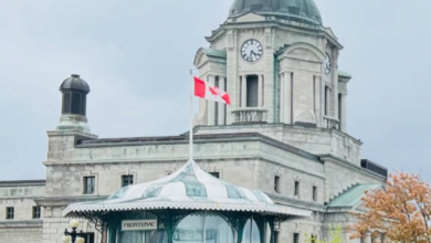 Historic stone building with a green-domed clock tower, Canadian flag, and a glass pavilion at the entrance; people seated nearby on benches.