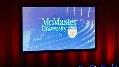Stage with a large screen showing the McMaster University logo and crest, framed by red curtains and blue chairs in front.