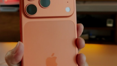 Person holding a rose-gold iPhone with a triple-camera module and Apple logo visible, indoors with a blurred screen in the background.
