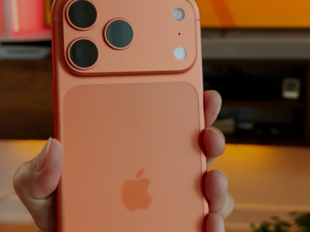 Person holding a rose-gold iPhone with a triple-camera module and Apple logo visible, indoors with a blurred screen in the background.