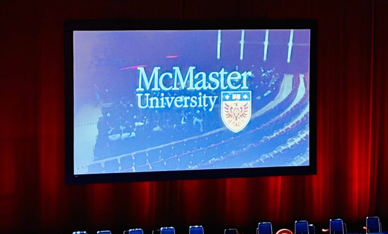 Stage with a large screen showing the McMaster University logo and crest, framed by red curtains and blue chairs in front.