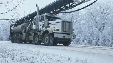 White heavy-duty aerial-ladder truck parked on a snow-covered road with a long extended boom and bucket at the end, winter trees in the background.