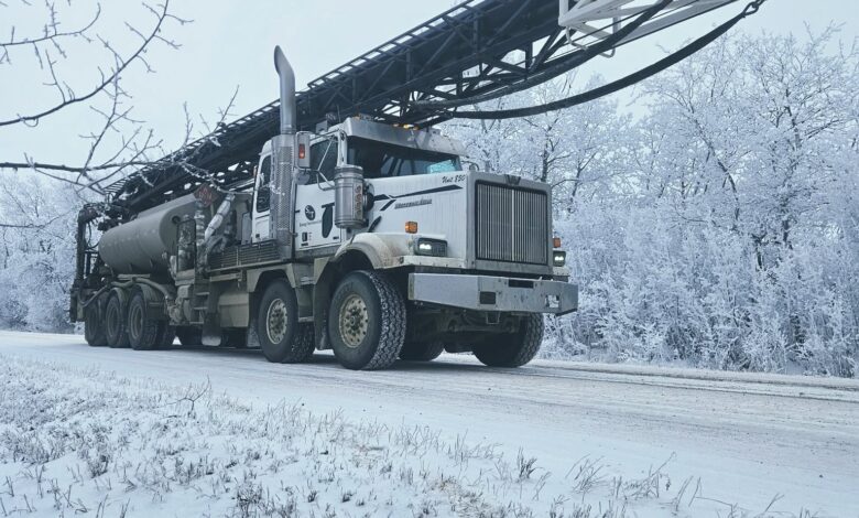 White heavy-duty aerial-ladder truck parked on a snow-covered road with a long extended boom and bucket at the end, winter trees in the background.