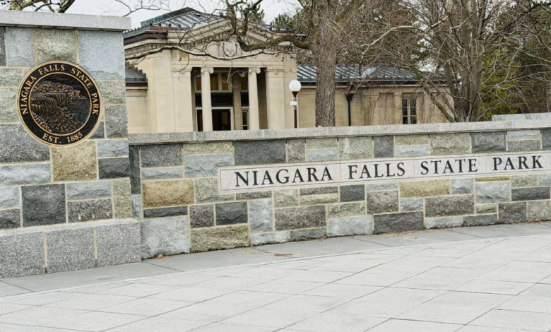 Stone entrance wall reading 'Niagara Falls State Park' with a circular park seal on the left and a neoclassical building beyond, leafless trees in view.