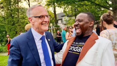 Two men in a park-like setting share a laugh at an outdoor event; one wears a blue suit and glasses, the other a cream-colored blazer with a brown lapel, surrounded by guests.