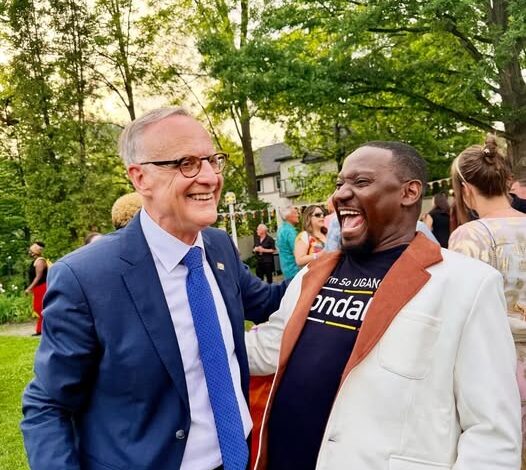 Two men in a park-like setting share a laugh at an outdoor event; one wears a blue suit and glasses, the other a cream-colored blazer with a brown lapel, surrounded by guests.