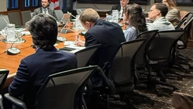 Professionals in a boardroom around a long conference table with laptops and papers; a Canadian flag and a large wall screen behind them.