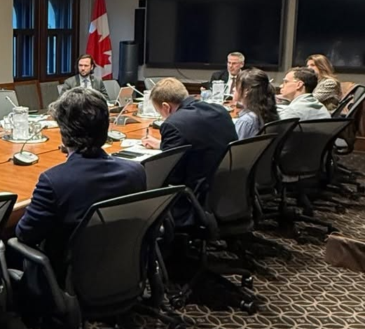 Professionals in a boardroom around a long conference table with laptops and papers; a Canadian flag and a large wall screen behind them.