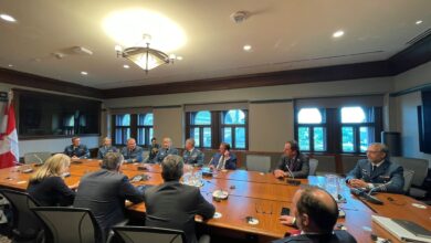 Group of people in military uniforms seated around a large wooden conference table in a formal meeting room with windows and a Canadian flag nearby, during daytime.