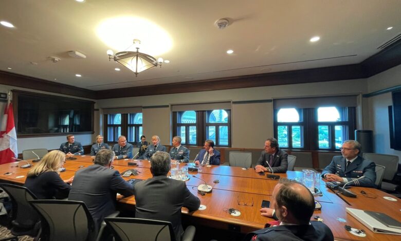 Group of people in military uniforms seated around a large wooden conference table in a formal meeting room with windows and a Canadian flag nearby, during daytime.