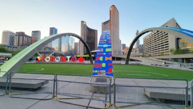 Public square in Toronto with large curved metal arches, a tall colorful sculpture, and the Toronto sign against a clear blue sky.