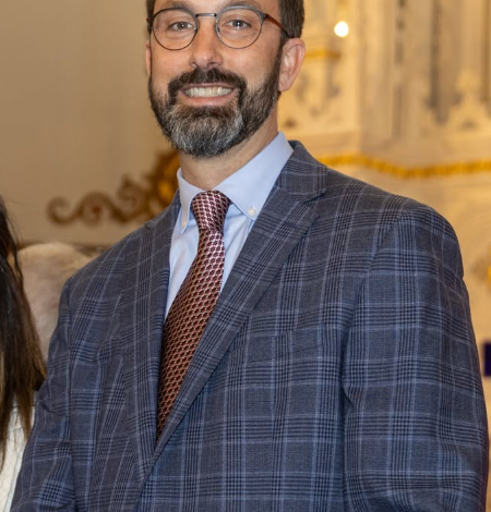 Smiling man in a blue checkered suit and brown tie at a formal event, neutral backdrop with ornate decor behind him.