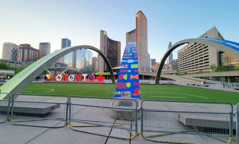 Public square in Toronto with large curved metal arches, a tall colorful sculpture, and the Toronto sign against a clear blue sky.