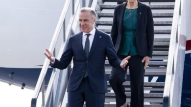 Two people in business attire walk down airplane stairs on the tarmac; the man in a dark suit gestures with open arms while the woman in a teal blouse and blazer follows.