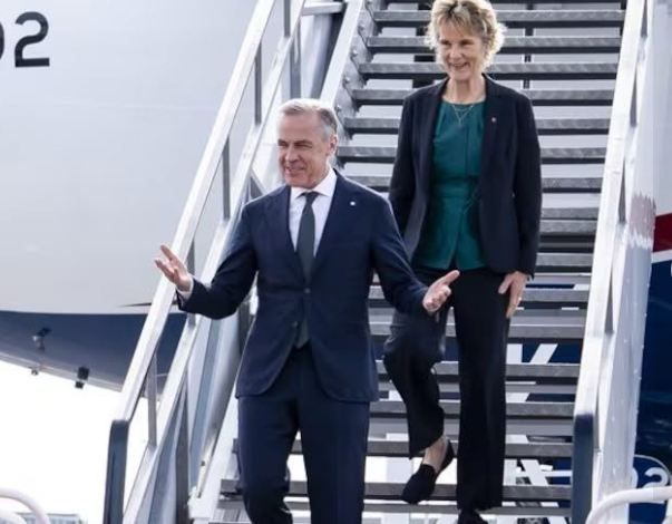 Two people in business attire walk down airplane stairs on the tarmac; the man in a dark suit gestures with open arms while the woman in a teal blouse and blazer follows.