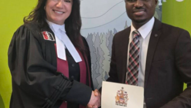 Two adults shake hands at a citizenship ceremony: a woman in academic robes and a man in a suit, holding a certificate with a red maple leaf; banner behind them displays '#MYCITIZENSHIP #MACITOYENNÉ' and Canada logos.