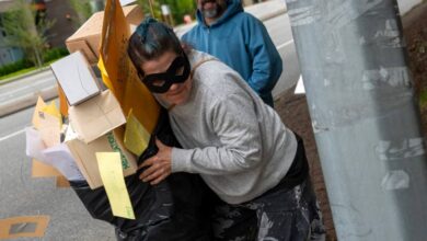 Person with a black mask and baggy clothes carrying a makeshift cardboard and paper display on a sidewalk; a sign behind reads 'YOUR MAIL YOUR SAY' and a man in a blue hoodie stands nearby.