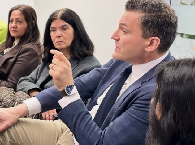 Man in a blue blazer speaks and gestures while seated with three colleagues in a business meeting.