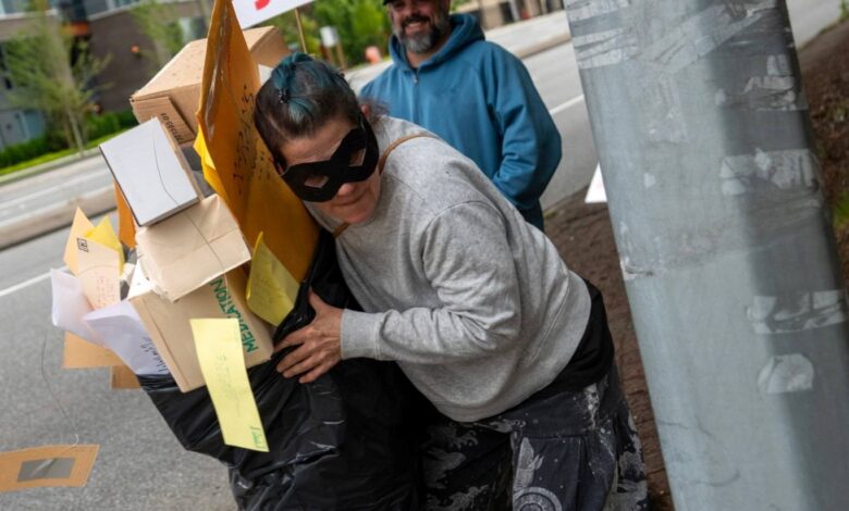 Person with a black mask and baggy clothes carrying a makeshift cardboard and paper display on a sidewalk; a sign behind reads 'YOUR MAIL YOUR SAY' and a man in a blue hoodie stands nearby.
