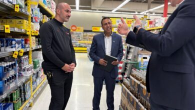 Three men stand in a supermarket aisle, two in dark uniforms and one in a suit, conversing near beer shelves with boxes on the right and left.