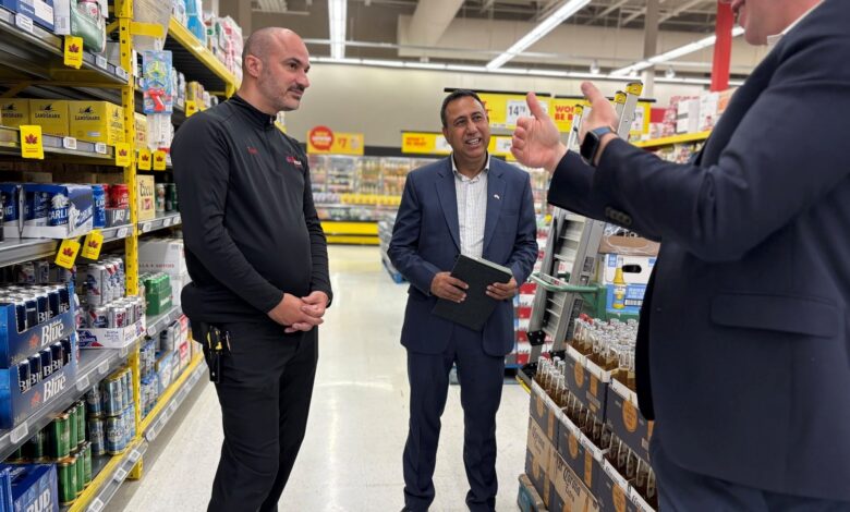 Three men stand in a supermarket aisle, two in dark uniforms and one in a suit, conversing near beer shelves with boxes on the right and left.