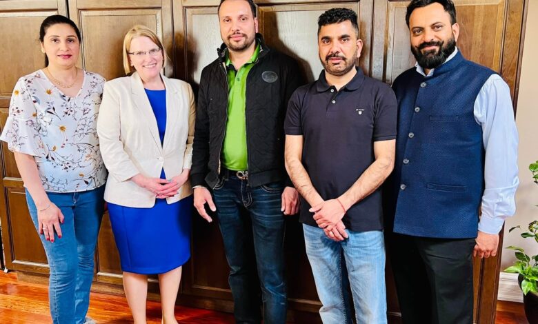 Group of five adults posing together indoors in front of wooden cabinets, dressed smart-c casual. All faces forward and smiling.