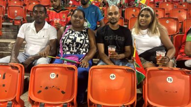 Group of four people sitting in stadium seats, drinks in hand, with orange seats around them and small flags nearby.