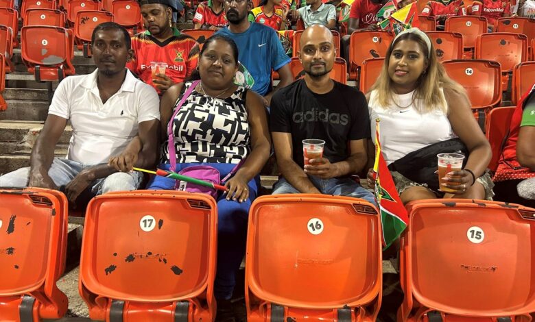 Group of four people sitting in stadium seats, drinks in hand, with orange seats around them and small flags nearby.