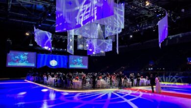 Formal gala in a dark arena with a circular, illuminated banquet platform, guests seated at round tables around a central stage, blue lighting overhead.