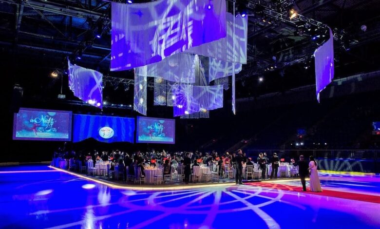 Formal gala in a dark arena with a circular, illuminated banquet platform, guests seated at round tables around a central stage, blue lighting overhead.