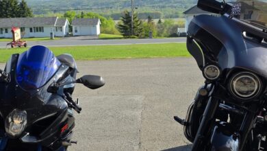 Two motorcycles parked facing each other in a driveway with a blue helmet nearby and rolling hills in the background.