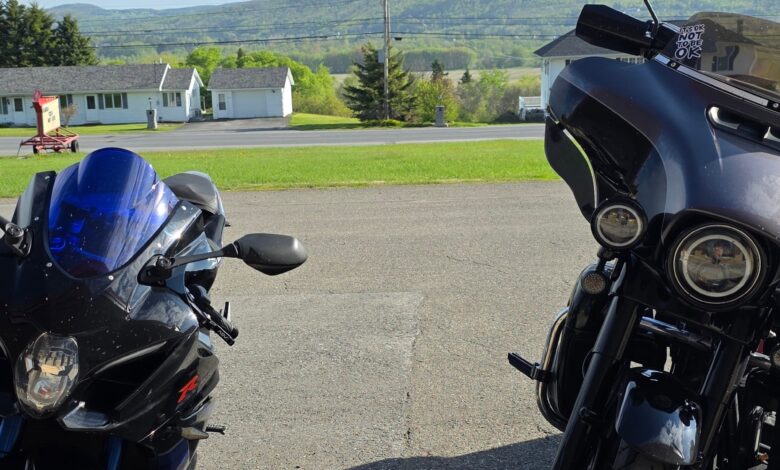 Two motorcycles parked facing each other in a driveway with a blue helmet nearby and rolling hills in the background.