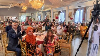 Banquet hall filled with guests at round tables, ornate chandeliers, draped ceiling, and a camera on a tripod in the foreground.