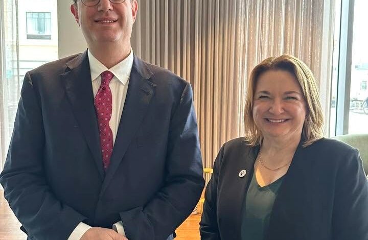 Two professionals indoors by a window, man in a navy suit and red tie, woman in a black blazer and green top, smiling.