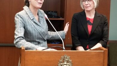 Two women at a wooden podium during a formal event; one speaks into a microphone while the other listens beside her.