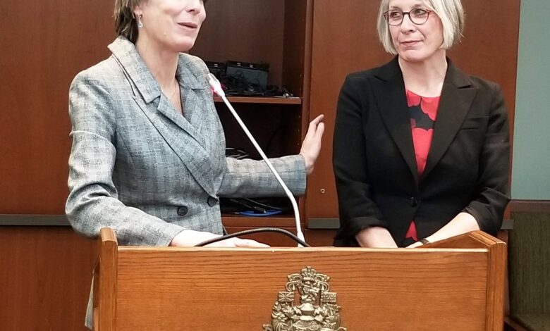 Two women at a wooden podium during a formal event; one speaks into a microphone while the other listens beside her.