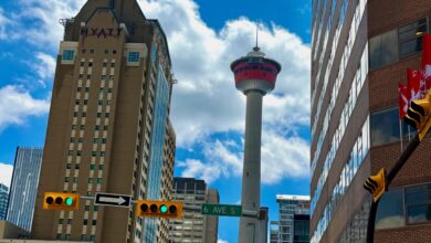 Urban street scene with tall beige and glass buildings, a distant tower, and a bright blue sky with clouds.