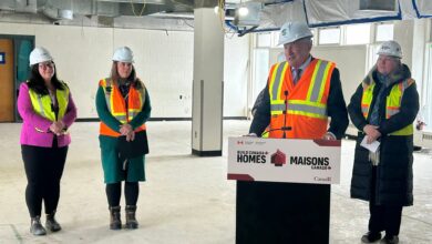 Group of four professionals in hard hats and high-visibility vests speaking at a construction site press event, with a podium reading Build Canada Homes/Maisons Canada.