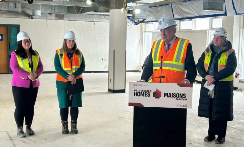 Group of four professionals in hard hats and high-visibility vests speaking at a construction site press event, with a podium reading Build Canada Homes/Maisons Canada.