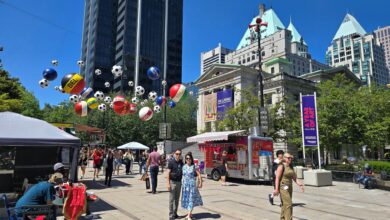 City square festival with hanging balloons, vendors, and people walking under a clear blue sky.