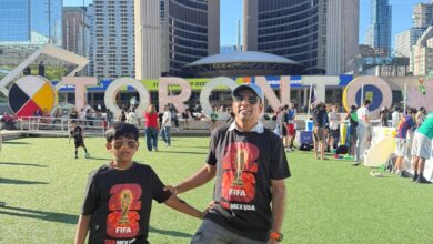 Two people in FIFA shirts posing with a colorful soccer ball on artificial turf, city skyline with curved glass towers in the background.