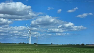 Wind turbines standing in a flat green field under a bright blue sky with scattered clouds.