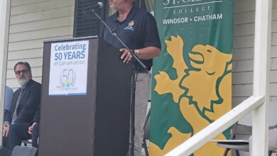 Man speaking at a podium on a porch, microphone in front, with a St. Clair College Windsor-Chatham banner nearby.