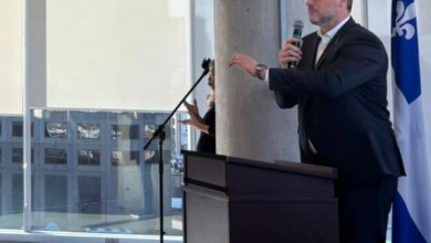 Man in a suit delivers a speech at a dark podium with a handheld microphone in a bright conference room; a Quebec flag is visible in the background.