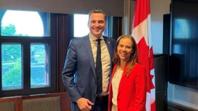 A man in a blue suit and a woman in a red blazer stand together in a meeting room with a Canadian flag in the background, smiling at the camera.