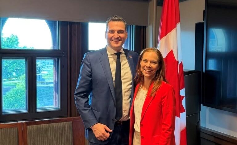 A man in a blue suit and a woman in a red blazer stand together in a meeting room with a Canadian flag in the background, smiling at the camera.